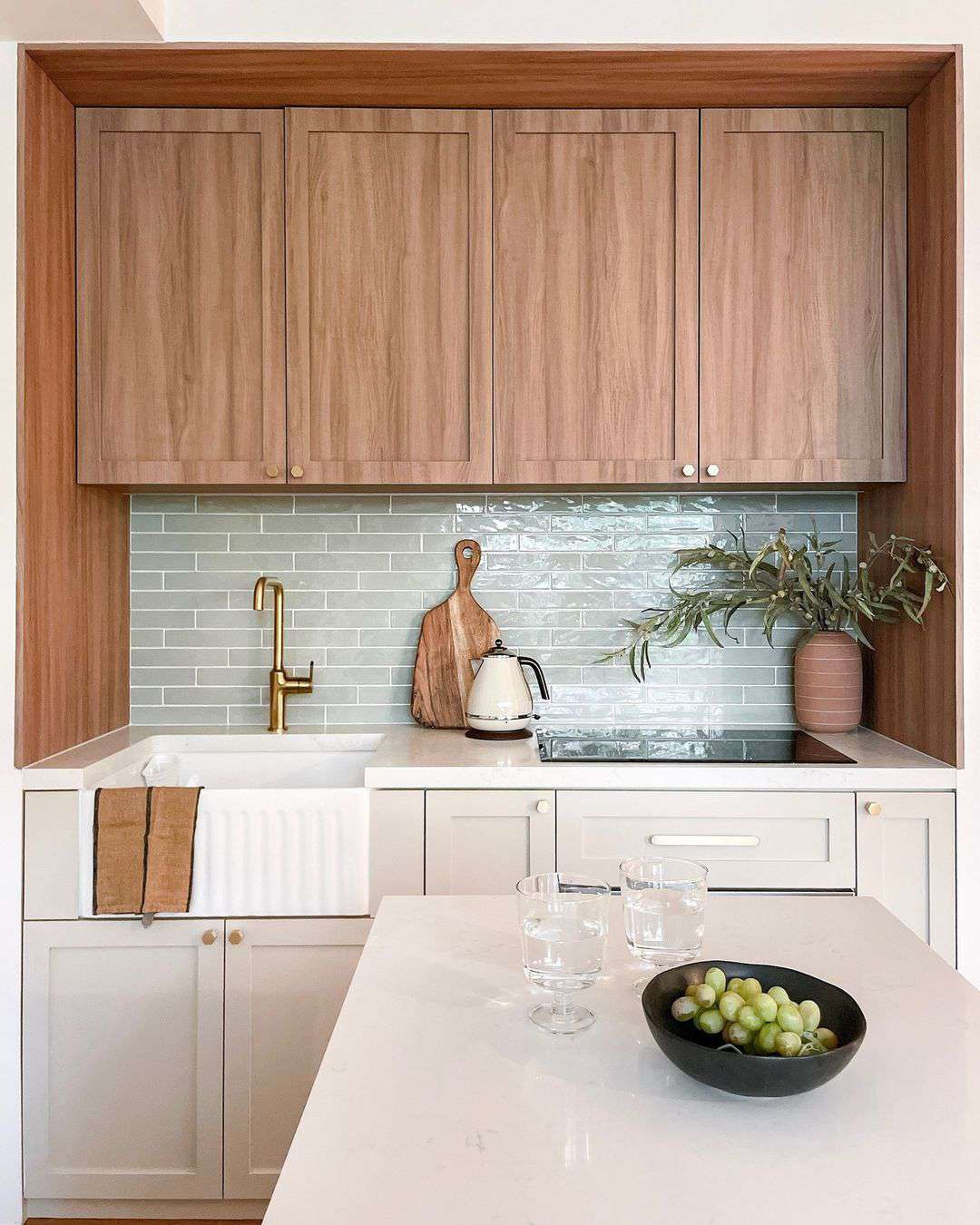 A neutral kitchen with warm wooden cabinets, a sage green backsplash, and gold accents