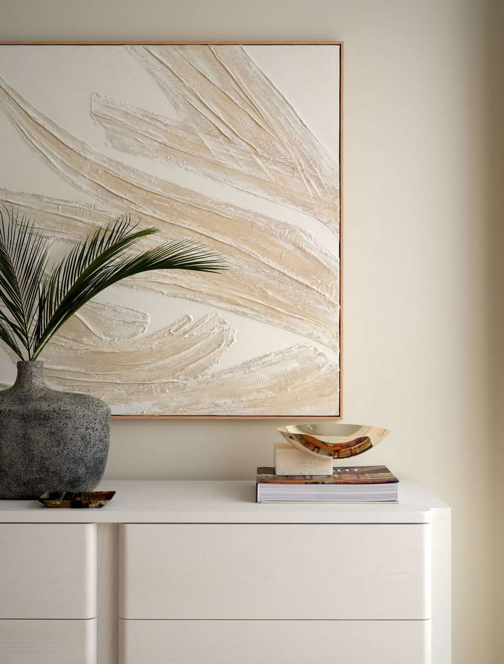 A textured abstract canvas above a white console table with a gold metal bowl, stone vase, and coffee table book on top