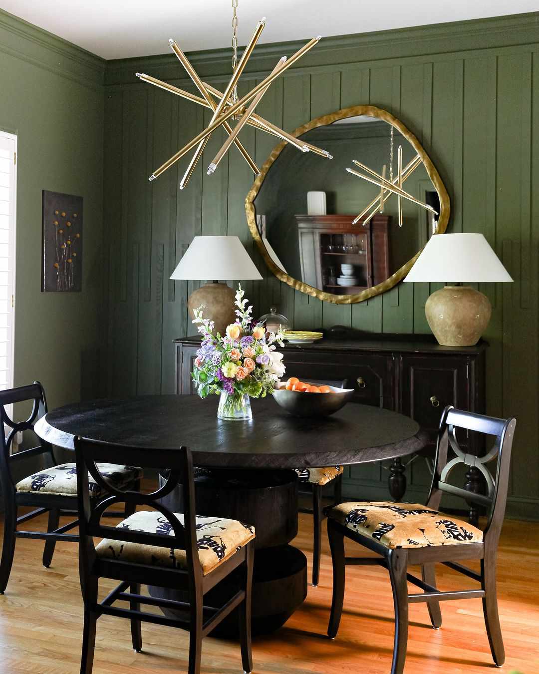 Moss green dining room with paneling, modern starburst brass chandelier, organic modern brass mirror, urn lamps, and black wood dining set